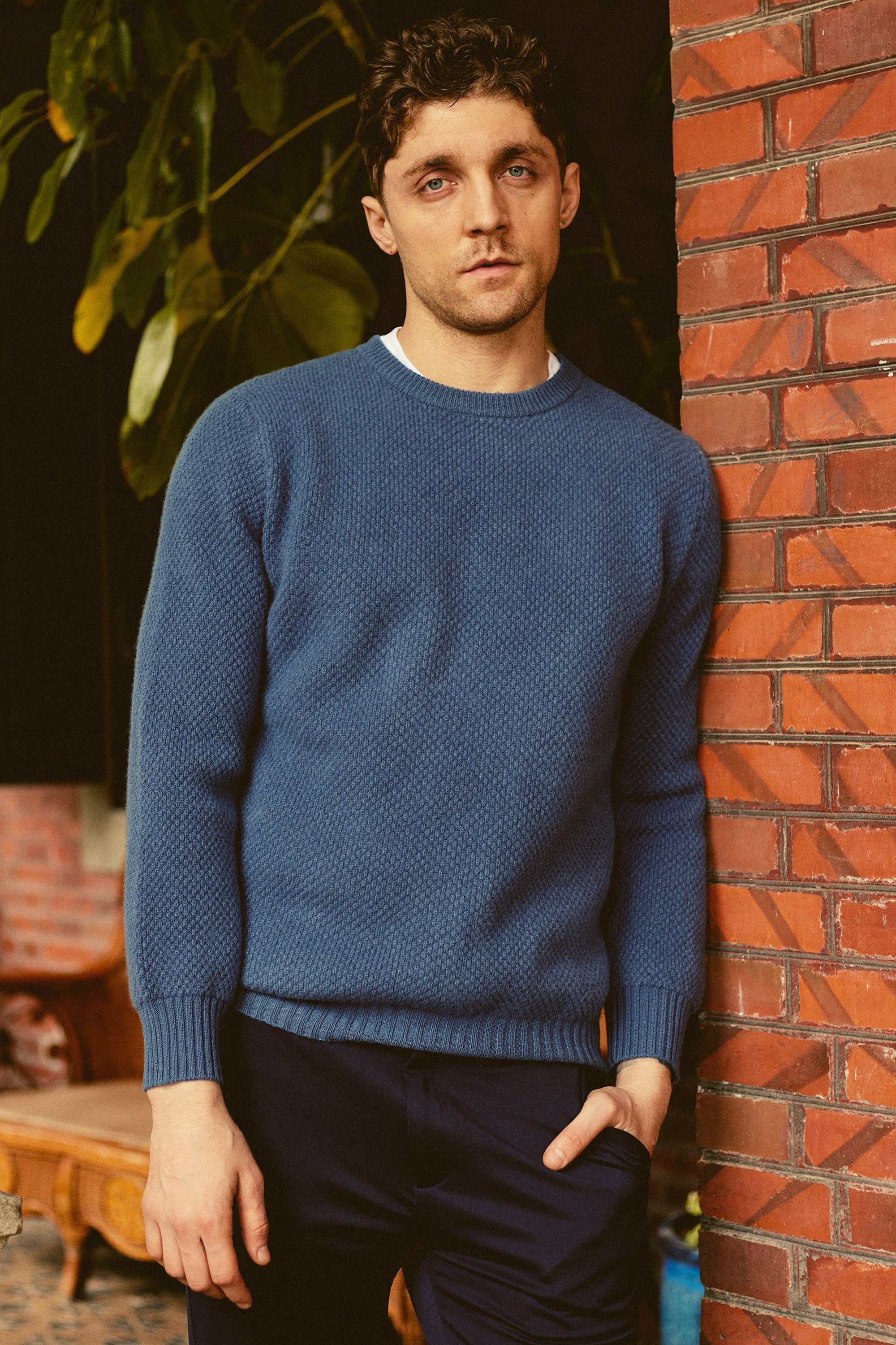 a man leaning against a brick wall wearing a blue sweater